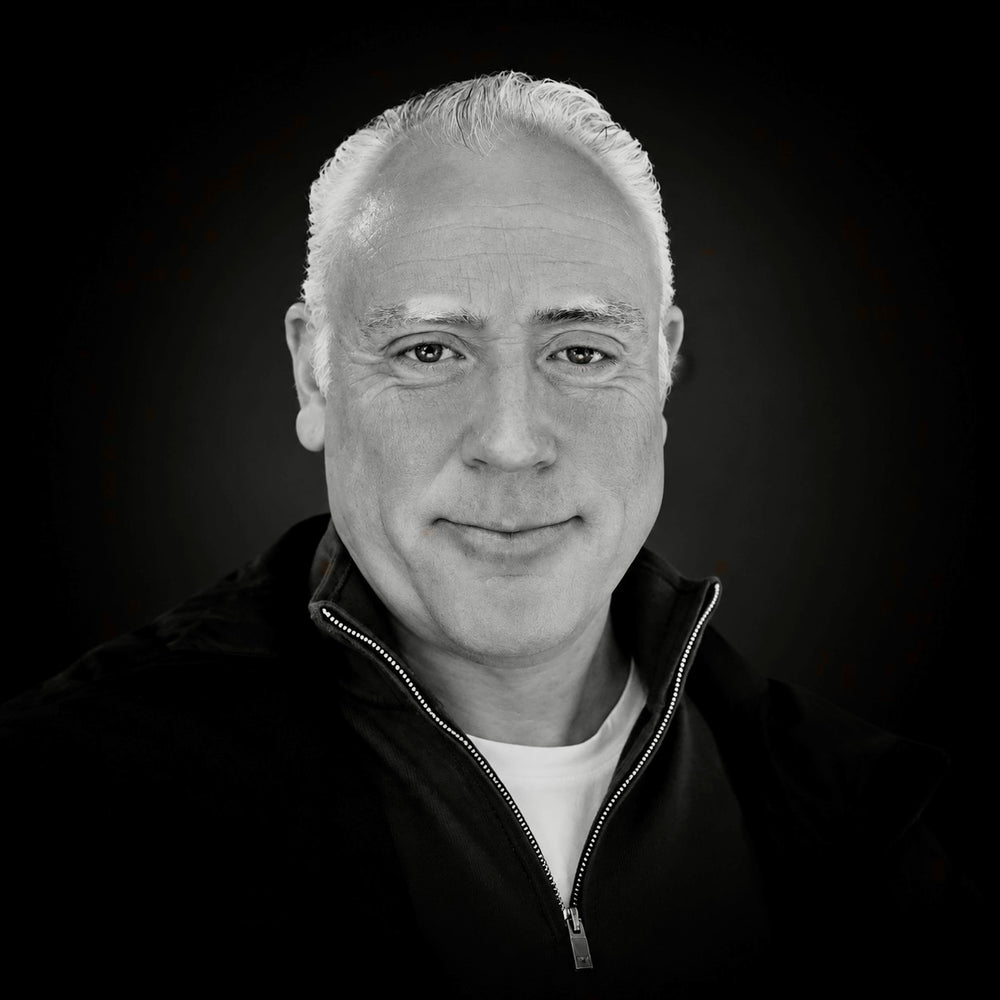 A black and white portrait of a man with short white hair, wearing a dark zip-up jacket over a white shirt, set against a dark background.