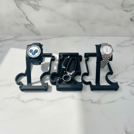 A black watch and accessory organizer stand , shown holding a black watch, a beaded bracelet, and a silver watch on a white marble surface.
