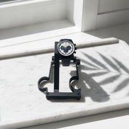 A black chronograph watch with a white dial and black mesh strap is displayed on a black stand on a white marble surface near a window, with palm leaf shadows cast across it.