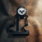 A black chronograph wristwatch with a white dial and black bezel, displayed on a black stand against a textured brown background.