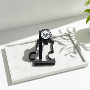 A black analog watch with a white dial and black mesh strap is displayed on a black stand placed on a white marble tray. The scene is lit by bright natural sunlight casting leaf shadows, with a green plant leaf visible in the upper right corner.