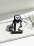 A black analog watch with a white dial and black mesh strap is displayed on a black stand placed on a white marble tray. The scene is lit by bright natural sunlight casting leaf shadows, with a green plant leaf visible in the upper right corner.