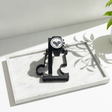 A black analog watch with a white dial and black mesh strap is displayed on a black stand placed on a white marble tray. The scene is lit by bright natural sunlight casting leaf shadows, with a green plant leaf visible in the upper right corner.