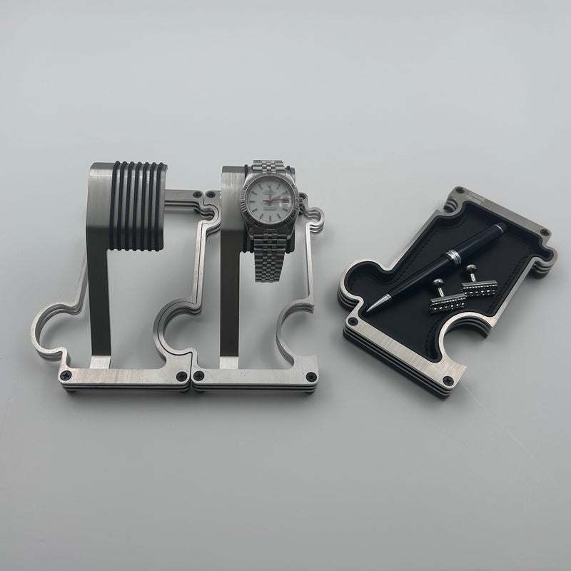 2 steel watch stands interlinked. The stand on the right has a white dial Rolex. Vanity tray on far right of image has a pen and cufflinks. white background