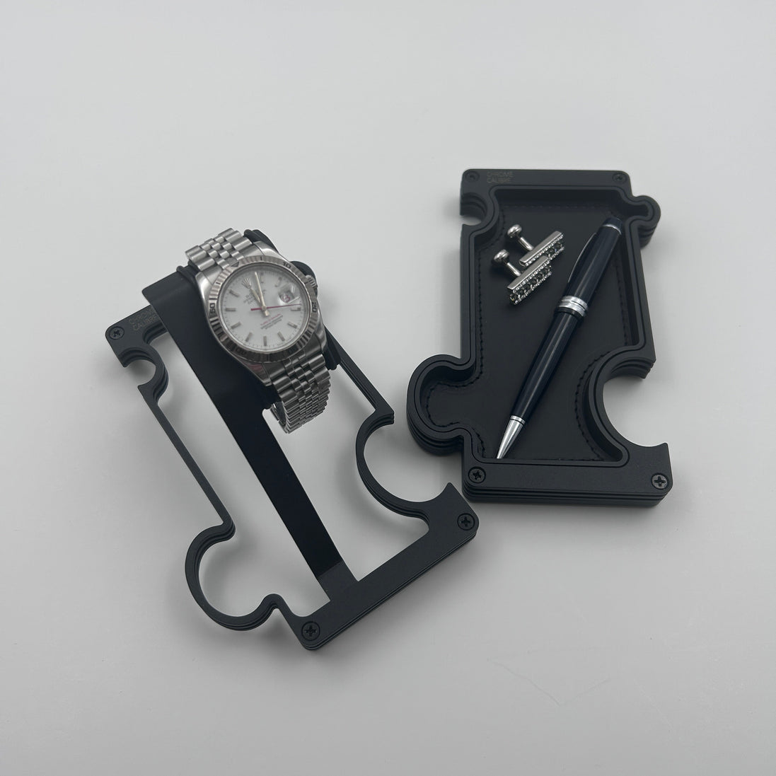 A black watch stand with white dial rolex sitting on it beside a vanity tray in the shape of a puzzle piece with a pen and cuff links resting on it