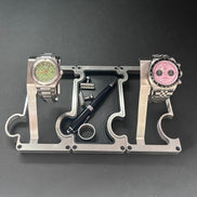 A silver metal watch stand with two slots holding stainless steel wristwatches, one with a green dial and one with a pink dial, alongside a black pen and a small ring on a dark gray background.