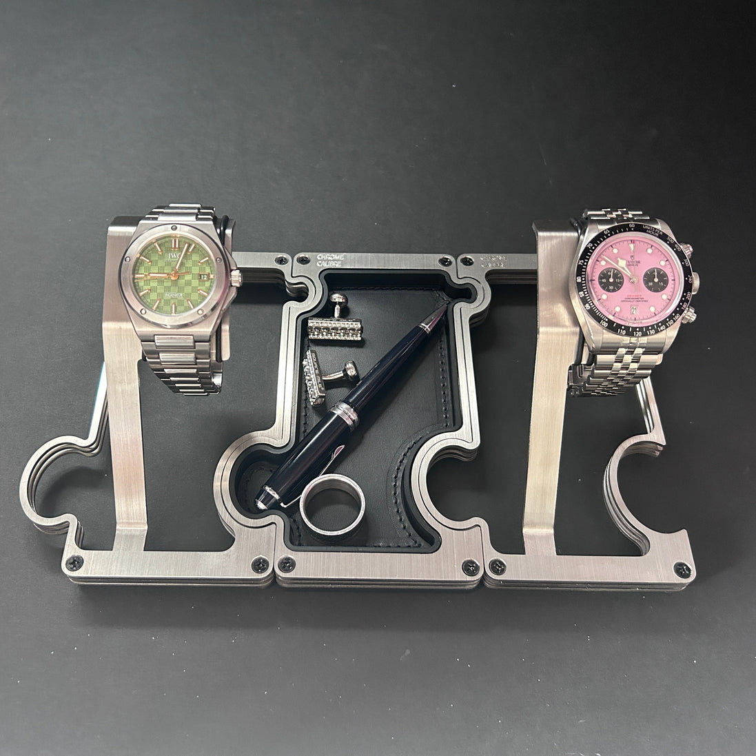 A silver metal watch stand with two slots holding stainless steel wristwatches, one with a green dial and one with a pink dial, alongside a black pen and a small ring on a dark gray background.