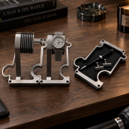 A silver metal watch display stand holding a wristwatch, shown on a wooden desk alongside a matching metal pen tray with a pen and small accessories. Books, a candle, and a plant are visible in the styled setting.