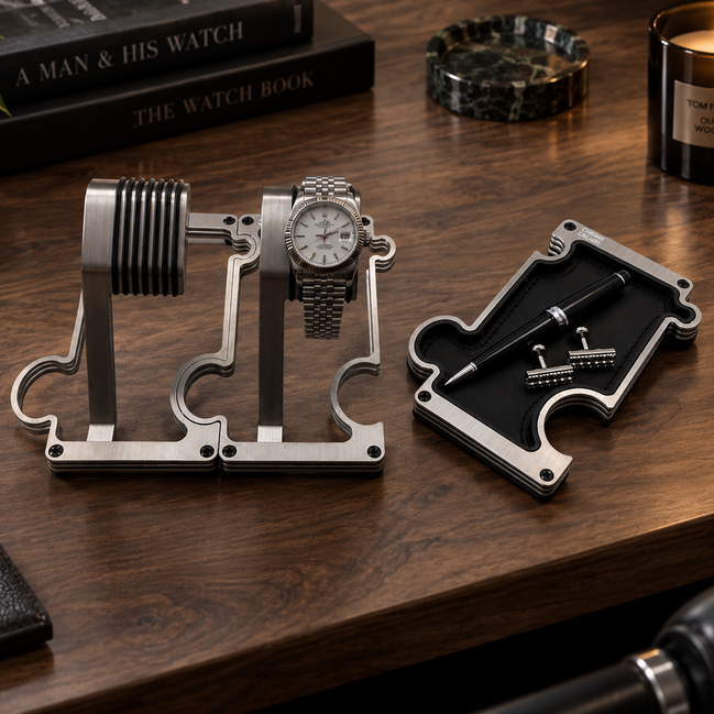 A silver metal watch display stand holding a wristwatch, shown on a wooden desk alongside a matching metal pen tray with a pen and small accessories. Books, a candle, and a plant are visible in the styled setting.