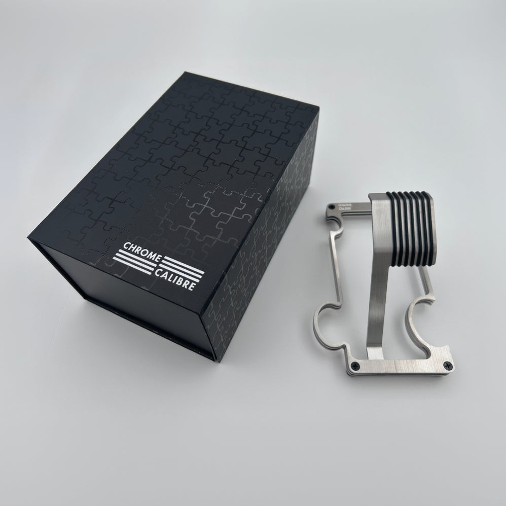 Metallic watch stand next to a black box with 'Chrome Calibre' branding on a light gray background