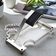 A brushed metal watch stand with a black ribbed rubber pad, shown on a light speckled countertop with a magazine, plant, and cup in the background.