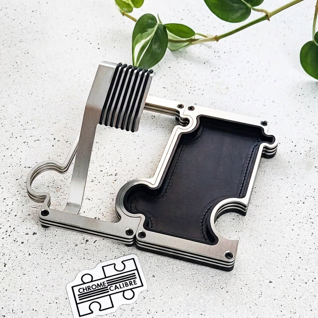 A brushed metal watch stand with a black leather insert, displayed on a light speckled surface with green plant leaves in the background and a 'Chrome Calibre' logo sticker at the bottom.
