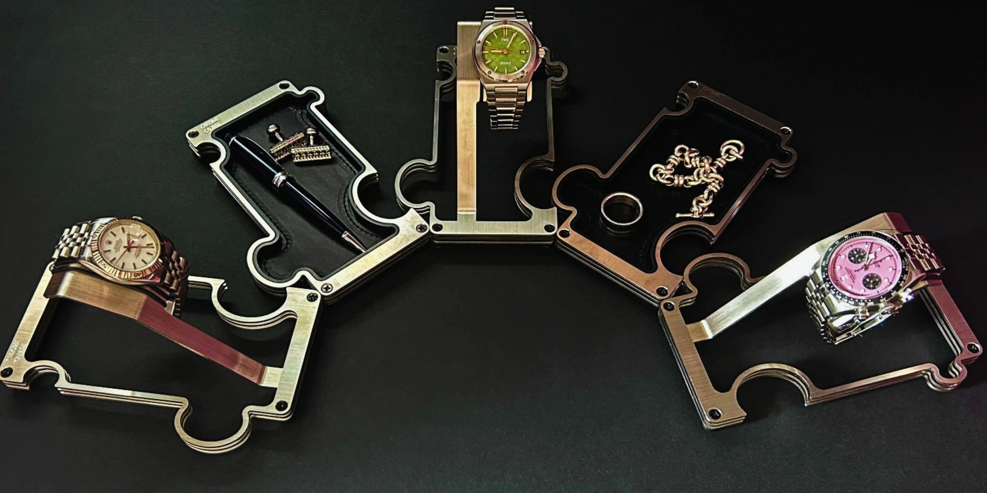 Four wristwatches displayed on a black background, each resting on a metallic puzzle-piece-shaped stand. The watches feature gold-tone and silver-tone cases with green, brown, and pink dials.