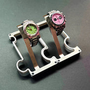 Two wristwatches displayed on a metallic watch stand against a dark gray background. The watch on the left has a green dial and pink bezel, while the watch on the right has a pink dial and dark bezel.