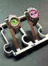 Two wristwatches displayed on a metallic watch stand against a dark gray background. The watch on the left has a green dial and pink bezel, while the watch on the right has a pink dial and dark bezel.
