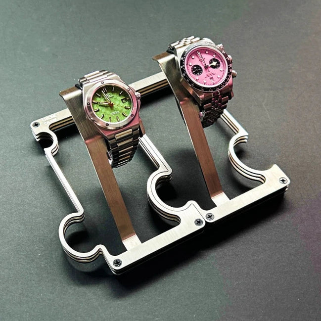 Two wristwatches displayed on a metallic watch stand against a dark gray background. The watch on the left has a green dial and pink bezel, while the watch on the right has a pink dial and dark bezel.
