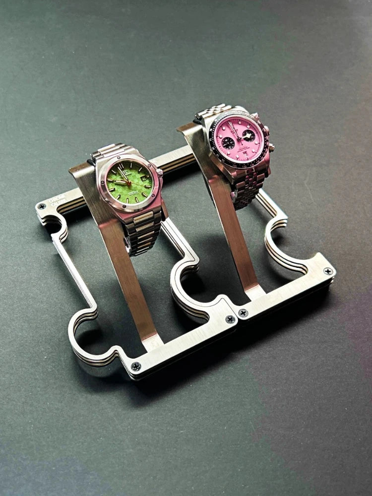 Two wristwatches displayed on a metallic watch stand against a dark gray background. The watch on the left has a green dial and pink bezel, while the watch on the right has a pink dial and dark bezel.