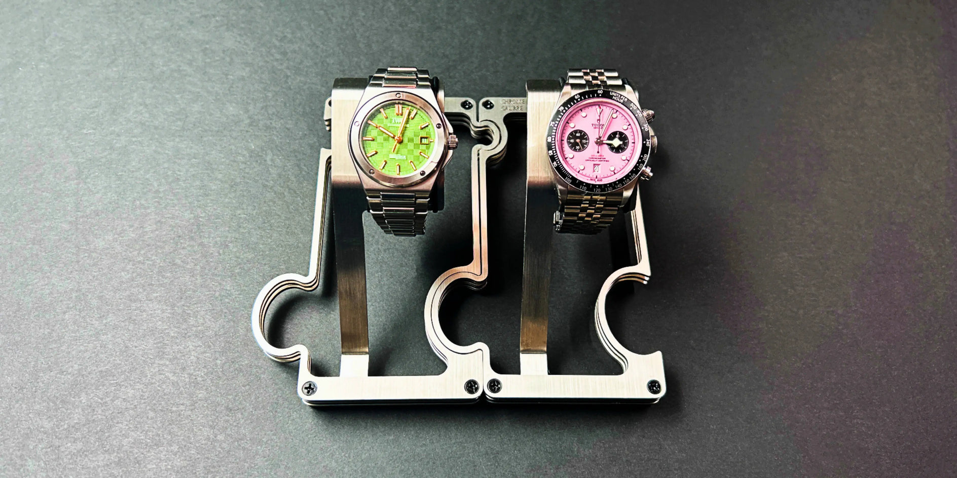 2 stainless steel watch stands on a grey background. the watch on the left has a green dial and the watch on the right has a pink dial