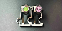 2 stainless steel watch stands on a grey background. the watch on the left has a green dial and the watch on the right has a pink dial