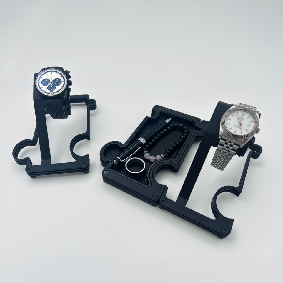 Two black watch stands with watches on a white background. a vanity tray connected to one of the watch stands and holding a pen, a ring and a beaded bracelet. the vanity tray has a leather insert
