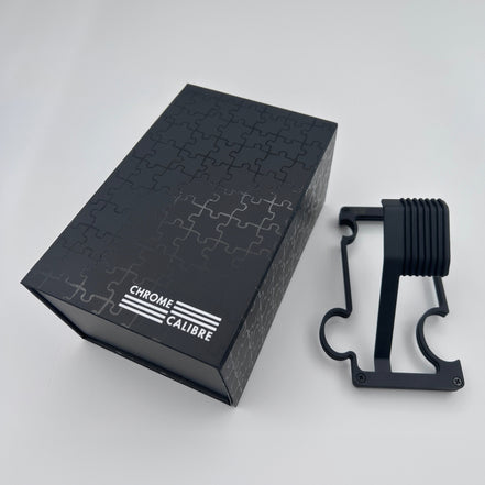 Black watch stand with 'Chrome Calibre' branding next to its packaging on a white background