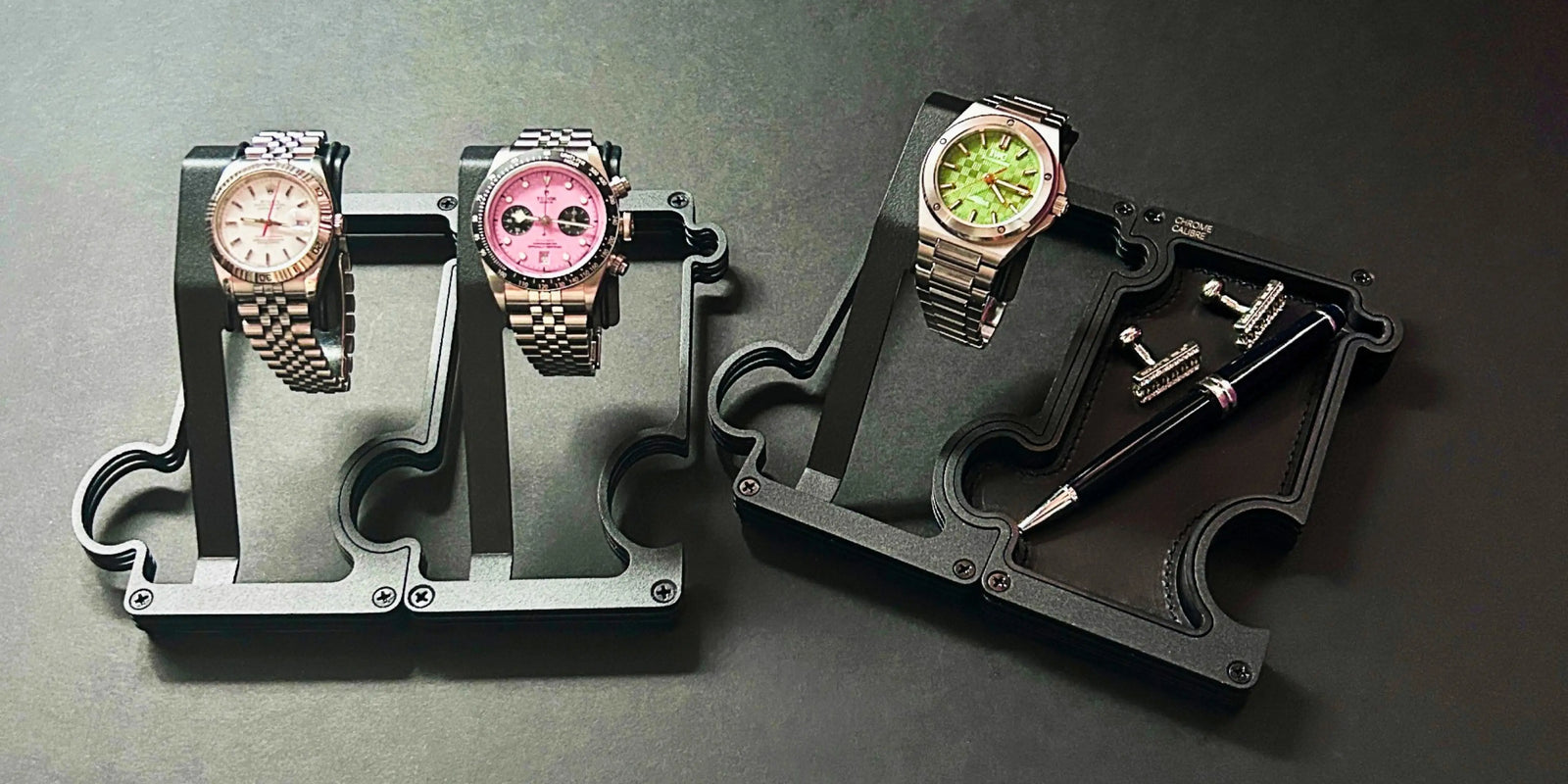 A pair of watch stands in black holding a rolex with a white dial and a tudor with a pink dial beside a watch stand with a vanity try attached. The vanity tray has a black pen and a pair of diamond cufflinks in it and the watch on the watch stand has an IWC with a green dial