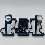 2 watches on 2 watch stands with a vanity tray in the middle holding a pen, a ring and a beaded bracelet. the watch stands are finished in black metal