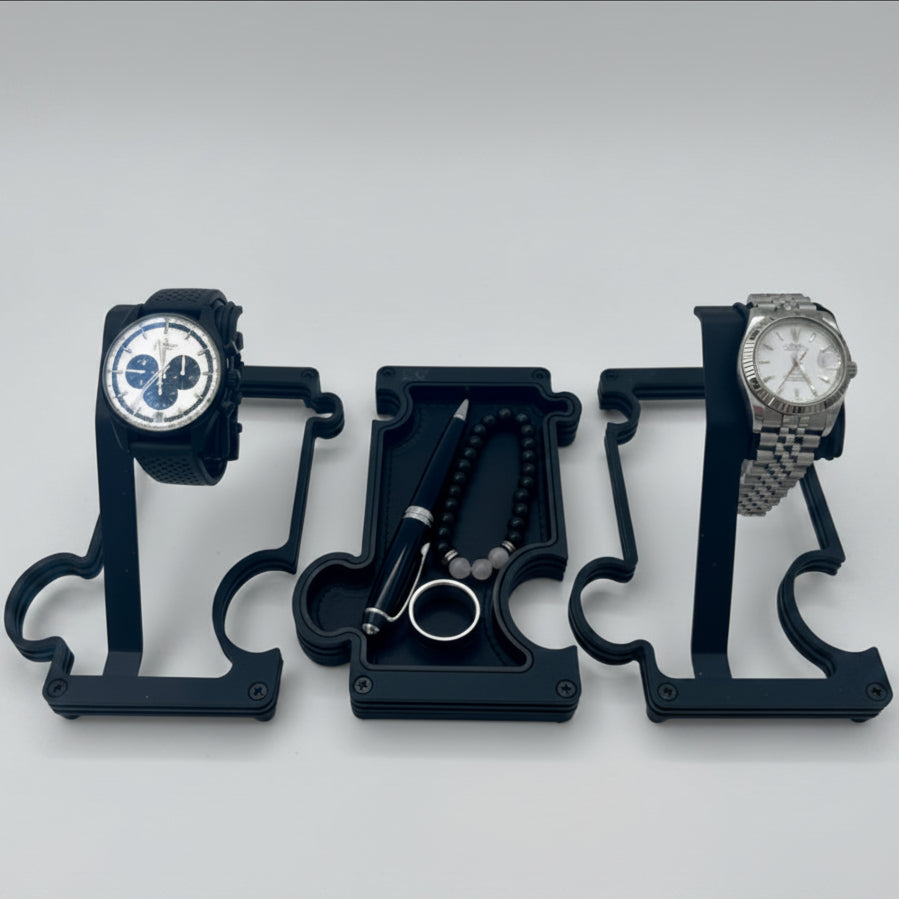 2 watches on 2 watch stands with a vanity tray in the middle holding a pen, a ring and a beaded bracelet. the watch stands are finished in black metal