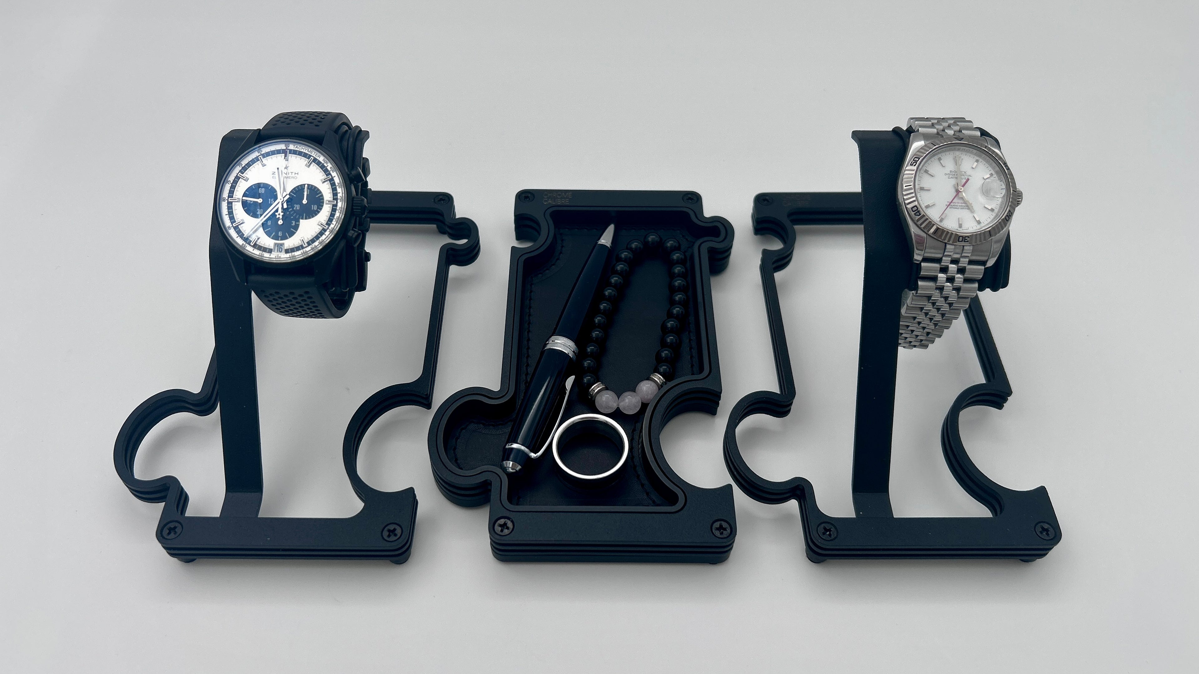 2 watches on 2 watch stands with a vanity tray in the middle holding a pen, a ring and a beaded bracelet. the watch stands are finished in black metal