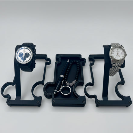 2 watches on 2 watch stands with a vanity tray in the middle holding a pen, a ring and a beaded bracelet. the watch stands are finished in black metal