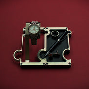 Watch and pen on a mechanical component against a red background. The product shown is a combination set of a watch display on the left interlinked with a valet tray on the right. The finish of this product is brushed steel with a silver colour. The watch shown on the watch holder is a Rolex Turn-O-Graph witha white dial and red second hand. The vanity tray to the right shows a leather insert in black real leather with a silver ring, silver cufflinks and a blakc and silver ball point pen