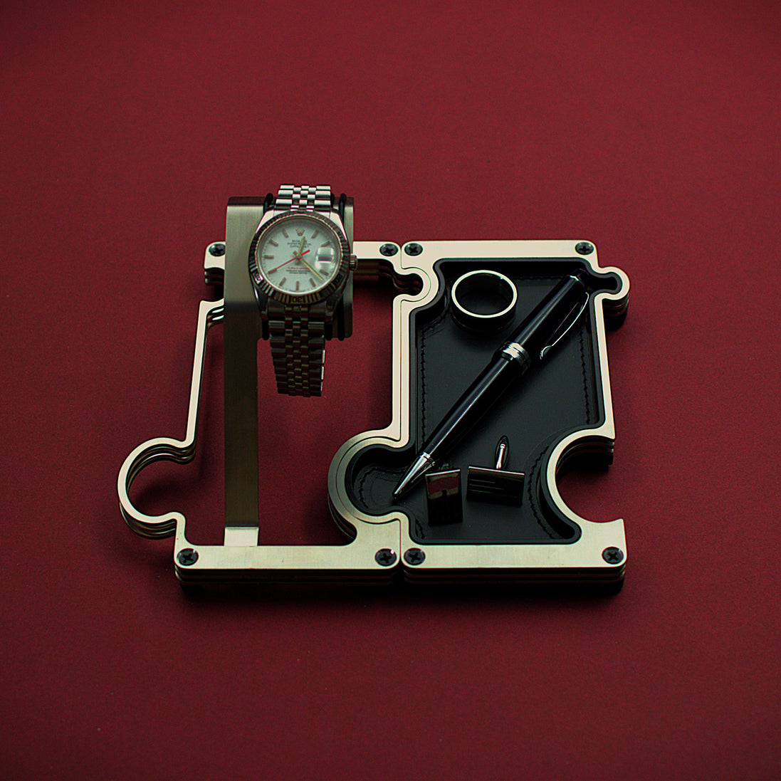 Watch and pen on a mechanical component against a red background. The product shown is a combination set of a watch display on the left interlinked with a valet tray on the right. The finish of this product is brushed steel with a silver colour. The watch shown on the watch holder is a Rolex Turn-O-Graph witha white dial and red second hand. The vanity tray to the right shows a leather insert in black real leather with a silver ring, silver cufflinks and a blakc and silver ball point pen