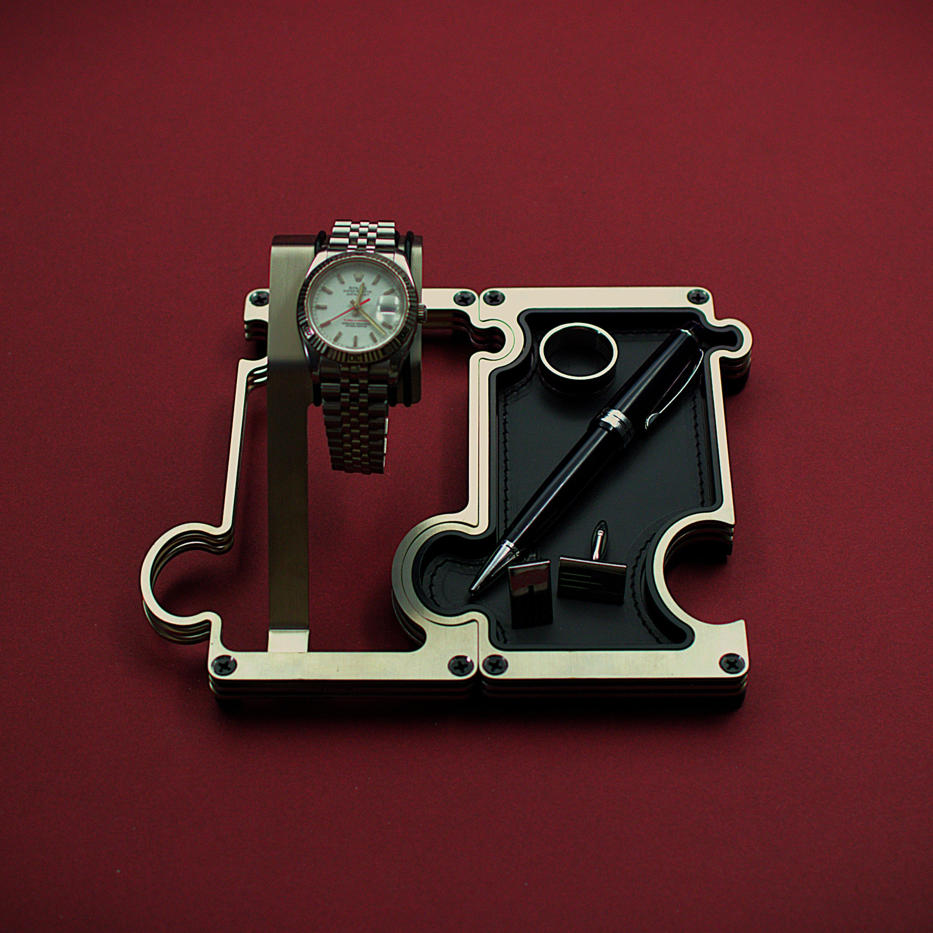 Watch and pen on a mechanical component against a red background. The product shown is a combination set of a watch display on the left interlinked with a valet tray on the right. The finish of this product is brushed steel with a silver colour. The watch shown on the watch holder is a Rolex Turn-O-Graph witha white dial and red second hand. The vanity tray to the right shows a leather insert in black real leather with a silver ring, silver cufflinks and a blakc and silver ball point pen