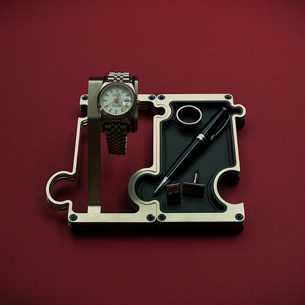 Watch and pen on a mechanical component against a red background. The product shown is a combination set of a watch display on the left interlinked with a valet tray on the right. The finish of this product is brushed steel with a silver colour. The watch shown on the watch holder is a Rolex Turn-O-Graph witha white dial and red second hand. The vanity tray to the right shows a leather insert in black real leather with a silver ring, silver cufflinks and a blakc and silver ball point pen