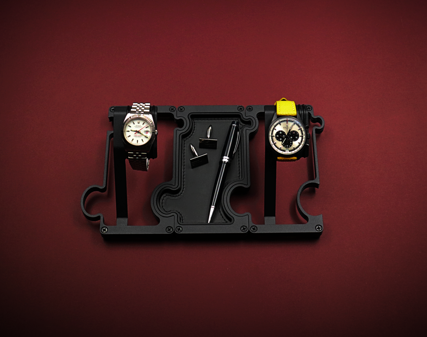 Black watch holder with two watches on a red background. The watch on the left is a white dial Rolex with red second hand. The watch on the right is a black ceramic Zenith El Primero with a panda dial. The black stand is made up of 2 watch holders and 1 valet tray. The valet tray has a leather insert and shows a pair of cufflinks and a black ballpoint pen