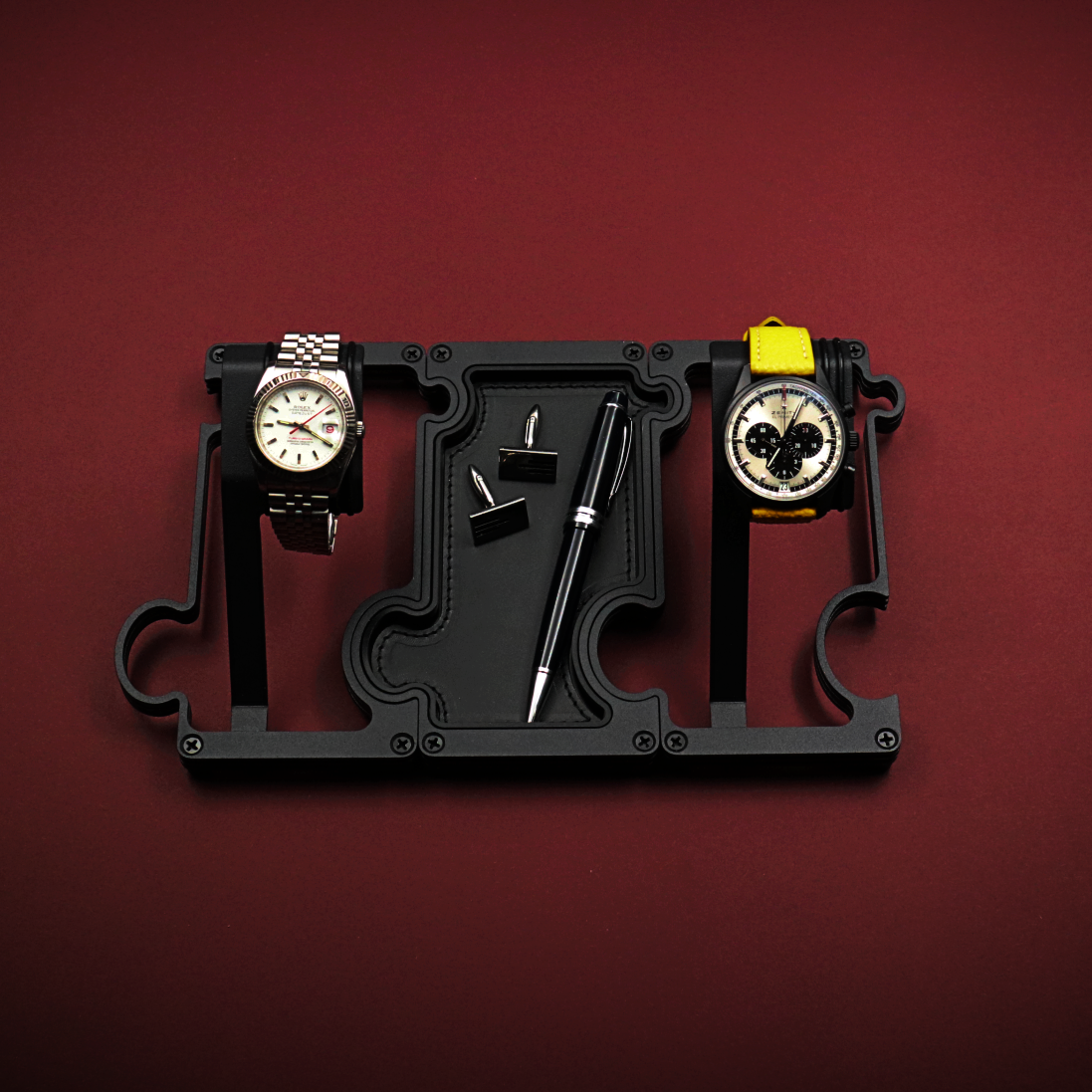 Black watch holder with two watches on a red background. The watch on the left is a white dial Rolex with red second hand. The watch on the right is a black ceramic Zenith El Primero with a panda dial. The black stand is made up of 2 watch holders and 1 valet tray. The valet tray has a leather insert and shows a pair of cufflinks and a black ballpoint pen