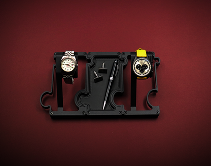 Black watch holder with two watches on a red background. The watch on the left is a white dial Rolex with red second hand. The watch on the right is a black ceramic Zenith El Primero with a panda dial. The black stand is made up of 2 watch holders and 1 valet tray. The valet tray has a leather insert and shows a pair of cufflinks and a black ballpoint pen