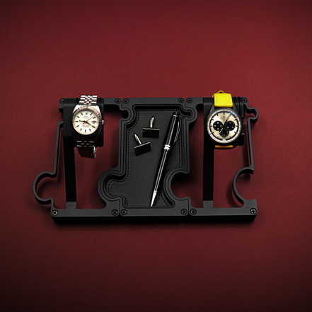 Black watch holder with two watches on a red background. The watch on the left is a white dial Rolex with red second hand. The watch on the right is a black ceramic Zenith El Primero with a panda dial. The black stand is made up of 2 watch holders and 1 valet tray. The valet tray has a leather insert and shows a pair of cufflinks and a black ballpoint pen