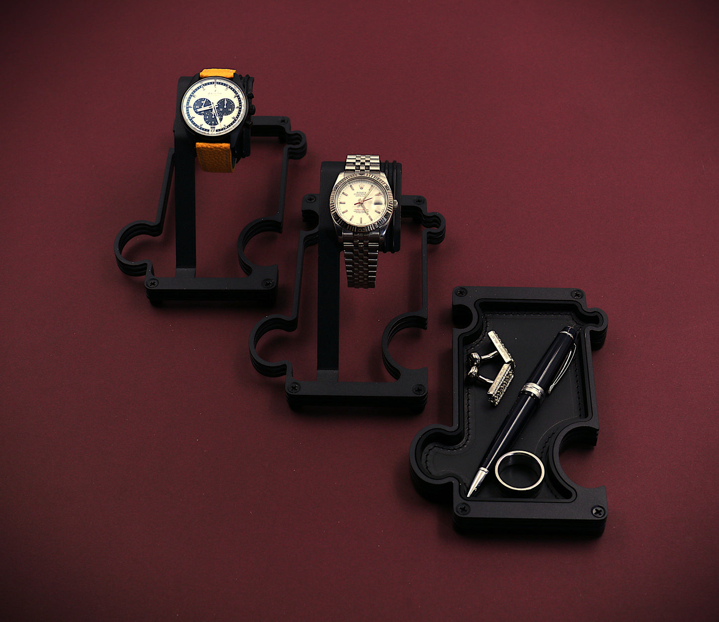 Three watches on black stands against a maroon background. The image shows a staggered selection of watch stands and a vanity tray. The finish of these products is a matt black steel. The watch stand to the top left of the image has a Zenith El Primero chronograph watch with a silver dial and black sub dials. It also has a yellow leather strap attached to the black watch case. The 2nd stand in the middle of the image has a white dial RolexTurn-O-Graph with a steel case and jubilee bracelet. The vanity tray