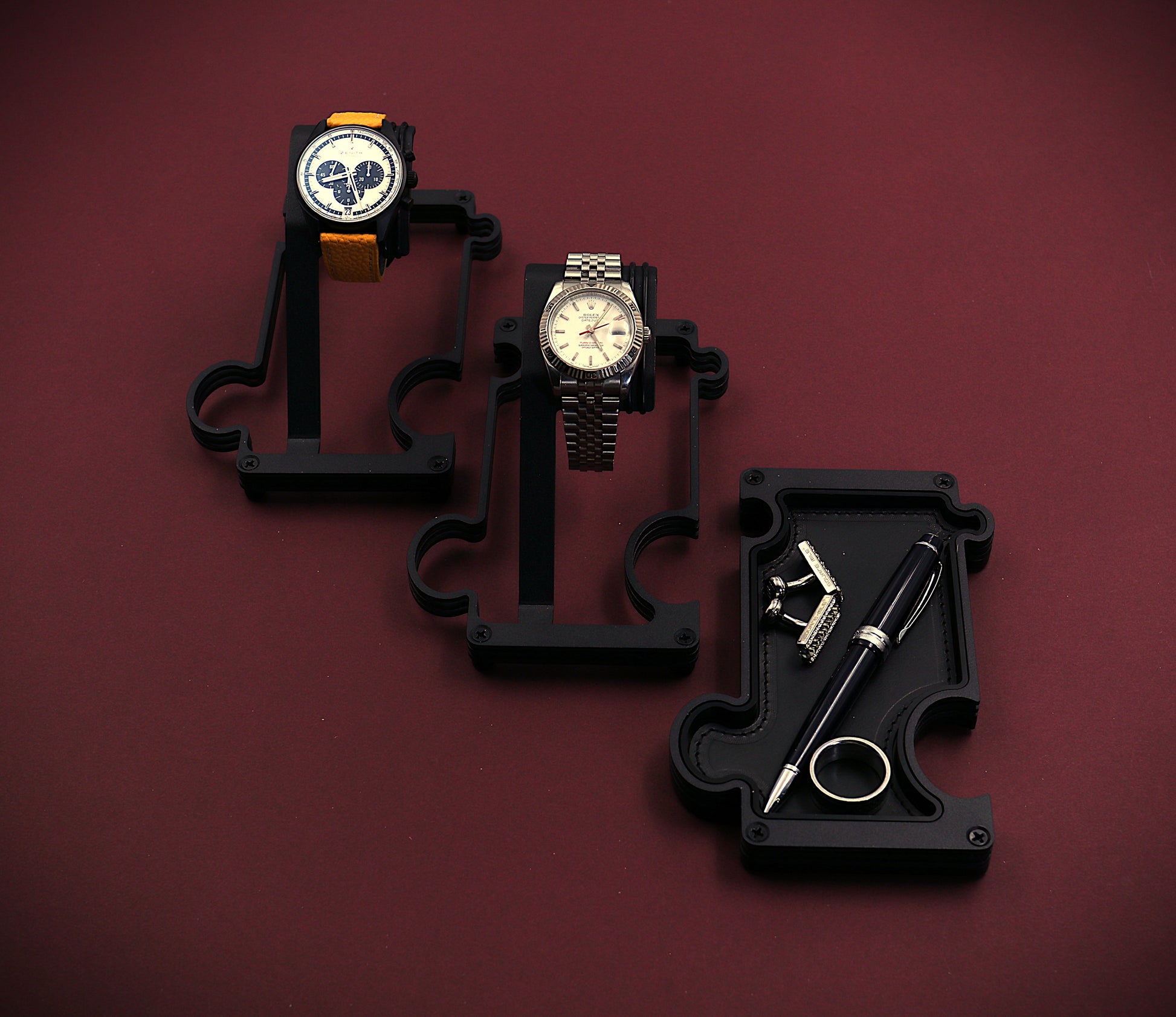 Three watches on black stands against a maroon background. The image shows a staggered selection of watch stands and a vanity tray. The finish of these products is a matt black steel. The watch stand to the top left of the image has a Zenith El Primero chronograph watch with a silver dial and black sub dials. It also has a yellow leather strap attached to the black watch case. The 2nd stand in the middle of the image has a white dial RolexTurn-O-Graph with a steel case and jubilee bracelet. The vanity tray