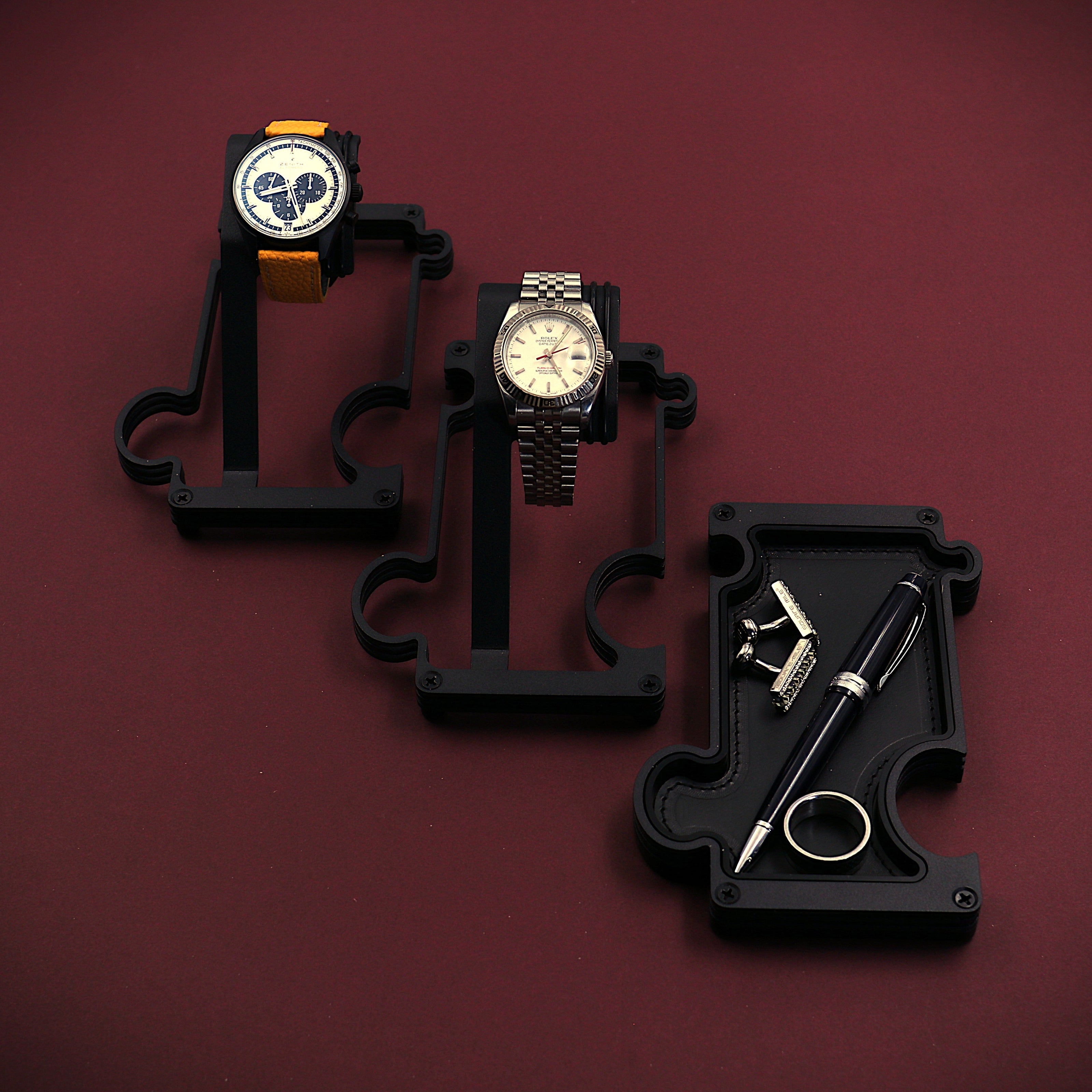 Three watches on black stands against a maroon background. The image shows a staggered selection of watch stands and a vanity tray. The finish of these products is a matt black steel. The watch stand to the top left of the image has a Zenith El Primero chronograph watch with a silver dial and black sub dials. It also has a yellow leather strap attached to the black watch case. The 2nd stand in the middle of the image has a white dial RolexTurn-O-Graph with a steel case and jubilee bracelet. The vanity tray