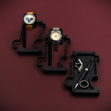 Three watches on black stands against a maroon background. The image shows a staggered selection of watch stands and a vanity tray. The finish of these products is a matt black steel. The watch stand to the top left of the image has a Zenith El Primero chronograph watch with a silver dial and black sub dials. It also has a yellow leather strap attached to the black watch case. The 2nd stand in the middle of the image has a white dial RolexTurn-O-Graph with a steel case and jubilee bracelet. The vanity tray