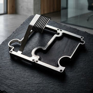 A watch stand and vanity tray set with a brushed silver metal frame and black textured ridges, resting on a dark slate surface in an indoor setting.