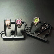 Two watch display stands on a dark gray surface, one holding two wristwatches (one white dial, one pink dial) and the other holding a watch with a green dial and a black pen and cufflinks