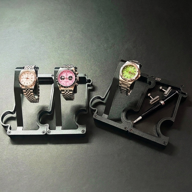 Two watch display stands on a dark gray surface, one holding two wristwatches (one white dial, one pink dial) and the other holding a watch with a green dial and a black pen and cufflinks