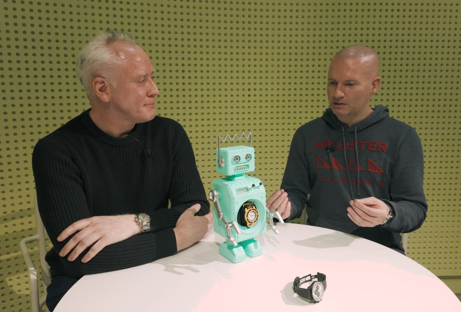 This image shows 2 people having a discussion as part of an interview for Scottish Watches Podcast. There is a green background and hey are seated at a table with the Retro-Bot watch winder in the centre of the table. 