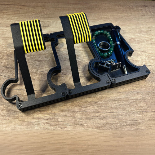 A black Metal Watch Stand Sett with yellow striped rubber pads, shown with its matching black storage tray and small hardware on a wooden surface.